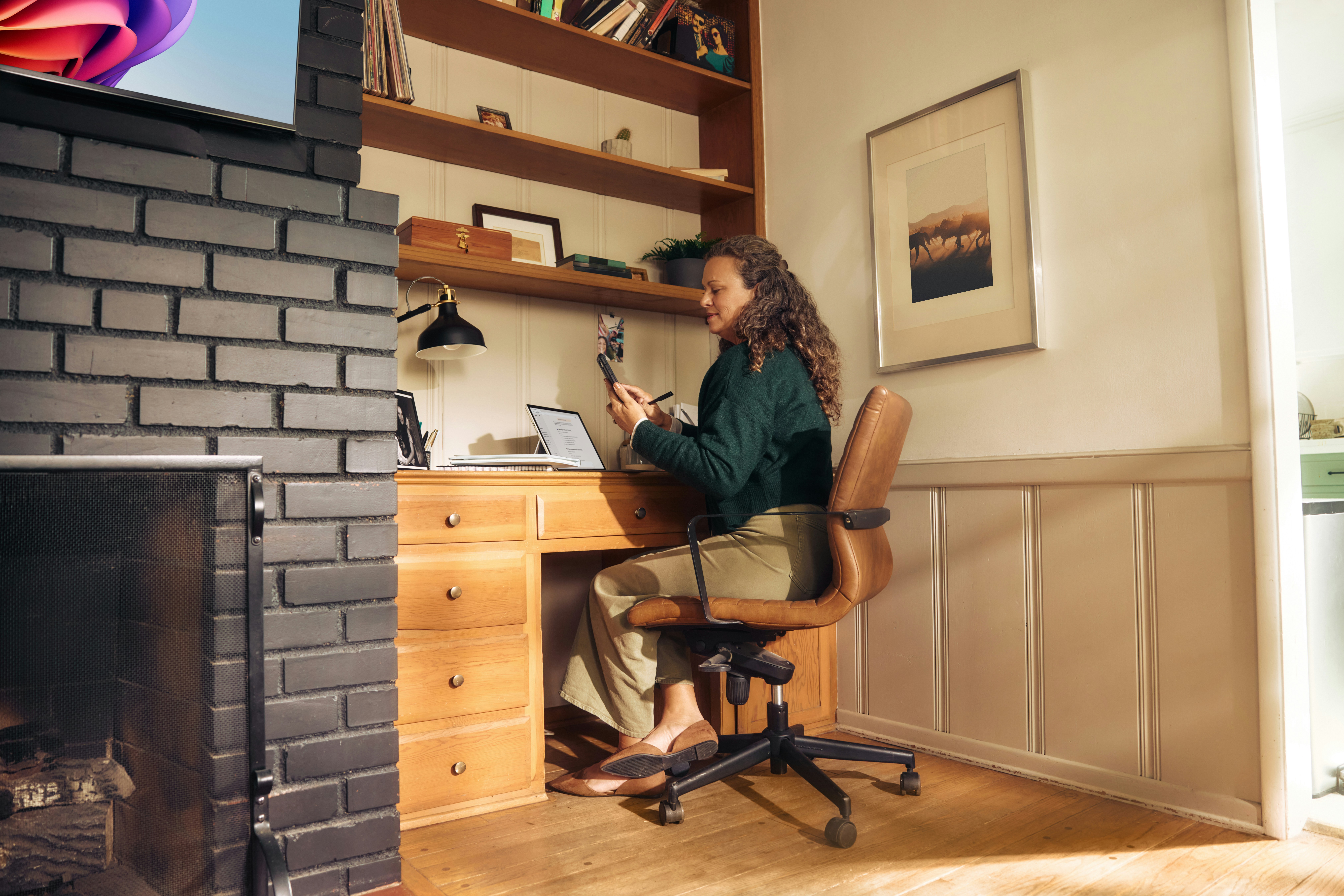 Woman sitting at a built-in desk nook using a stylus on her phone, with a laptop beside her, wooden shelves above, and a fireplace with a mounted TV to the left.