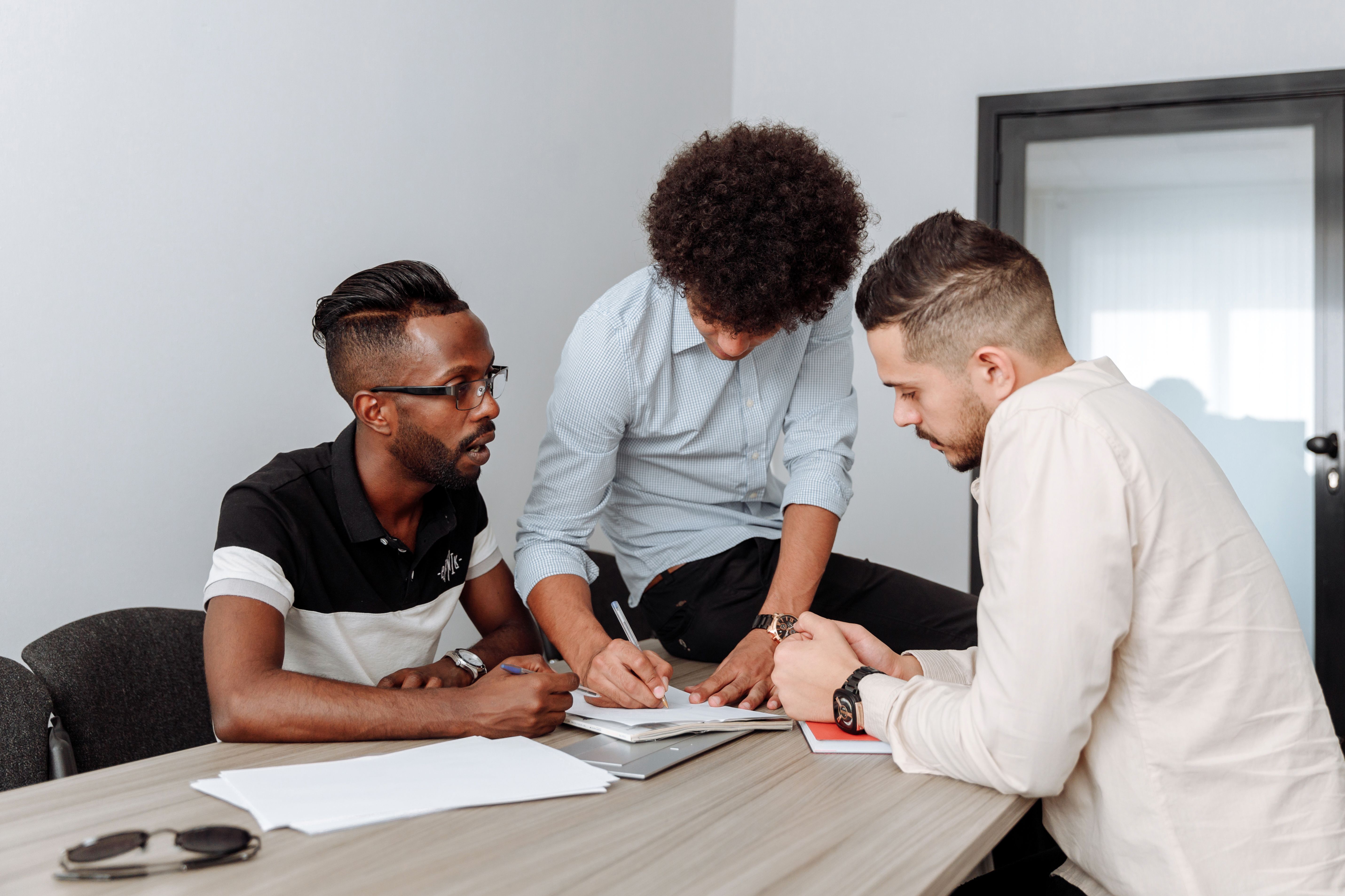 Three colleagues at a table, one writing in a notebook as the others discuss the document.
